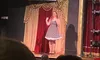A woman in a polka dot dress is standing on stage in front of a red and gold curtain with an audience watching, while a projected image of out-of-focus lights appears above her.