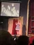 A woman in a polka dot dress is standing on stage in front of a red and gold curtain with an audience watching, while a projected image of out-of-focus lights appears above her.