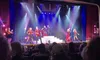 A group of performers in vibrant costumes entertains a seated audience on a brightly lit stage.