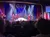 A group of performers in vibrant costumes entertains a seated audience on a brightly lit stage.