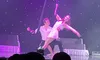 A pair of performers is engaged in a dance or aerial routine on stage, illuminated by purple lighting, with an audience watching in the foreground.