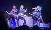 A group of performers energetically play drums on a brightly lit stage in front of an audience.