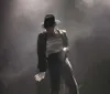 A performer in a glittery outfit sings energetically on stage under dramatic lighting, with an audience watching from below.