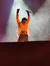 A performer on stage raises his arms triumphantly while holding a microphone, with dramatic lighting and smoke effects in the background.