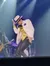A performer dressed in a white hat and striped jacket energetically dances on stage with a band in the background.