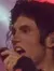 A performer on stage strikes a dynamic pose, illuminated by dramatic stage lighting with a vivid red screen in the background.