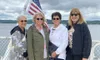 Four women wearing jackets and sunglasses stand together on a boat with an American flag in the background.