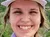 A smiling person wearing a white hat and a purple shirt takes a selfie with a scenic backdrop of forested hills and mountains under a cloudy sky.