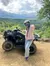 A person wearing a helmet and mask poses next to an ATV on a scenic dirt trail with a lush green landscape in the background.