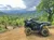A black ATV is parked on a dirt path overlooking a lush, green mountainous landscape under a cloudy sky.