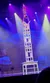 A performer balances on a tall stack of chairs while doing a handstand during a stage performance with colorful lighting.
