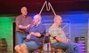 Three men are on stage, with two seated and wearing plungers on their heads while the third stands and interacts with them.