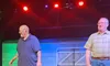 Two men are standing on a stage with chairs and barrels in the background, while one extends a hand towards another person off-frame.