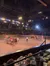 The image shows an indoor arena with an audience watching a performance featuring two horse-drawn wagons and a person standing between them on the sand-covered ground.
