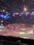 This image depicts a festive indoor arena show with performers in costumes riding on horses and pulling a sleigh, under artificial snow and colorful stage lights.