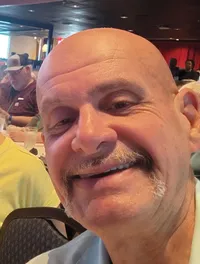 A man and woman are smiling for a selfie in a crowded indoor setting with tables and people in the background.