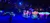 The image depicts a vibrant stage performance featuring large, colorful flowers and butterfly props under dramatic blue lighting.