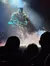 A large, illuminated figure dressed in a warrior costume stands on stage amidst smoke, viewed by an audience silhouetted in the foreground.