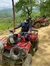 A person is sitting on a red ATV, smiling under a tree with a scenic view of mountains and a forest in the background.