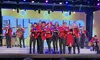 The image shows a group of musicians in red jackets performing energetically on a brightly lit stage with instruments, including violins and guitars.