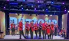 A group of performers in red jackets are playing violins on a brightly lit stage in front of an audience.