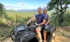 A couple is sitting on an ATV on a dirt path with a scenic view of a forested landscape and a blue sky with clouds in the background.