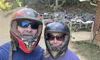 Two people wearing helmets and sunglasses pose for a selfie in front of parked all-terrain vehicles and a wooded area.