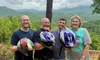 Four people with helmets stand smiling together on a muddy trail with a scenic forest and mountain backdrop.