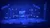 Three performers on a stage are surrounded by blue lighting and geometric structures.