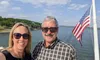 A man and a woman are smiling in front of a body of water with an American flag in the background.