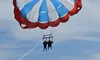 Two people are parasailing under a red, white, and blue parachute against a clear sky.