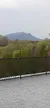The image shows a scenic view of a distant mountain under an overcast sky, with lush green trees in the foreground and a black metal railing in the midground.