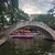 A stone arch bridge crosses over a river with a pink boat full of people, colorful umbrellas, and trees in the background.
