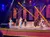 A lively musical performance is taking place on stage with a band and dancers under colorful stage lights.