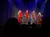 Four performers in red and black outfits are on stage under blue and white spotlights, entertaining an audience.