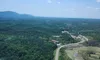 The image shows an aerial view of a scenic landscape with a winding road, lush greenery, and mountains under a clear blue sky with a few clouds.
