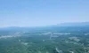 The image shows an aerial view of a vast, lush green landscape with distant mountains under a clear blue sky.