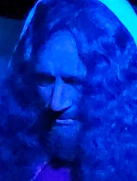 The image depicts a person with long hair and a beard, dressed in a white robe and purple cloak, kneeling and praying in a blue-lit setting surrounded by rocks and foliage.