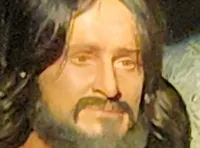 The image depicts a lifelike scene of a man in a white robe standing near a tomb entrance, with a seated woman beside it, both illuminated by spotlights against a dark background.