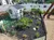 A small, fenced garden with various plants surrounds a miniature white house, with people and scenic views in the background.