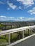 The image shows a scenic view of a wooden walkway with railings, set against a backdrop of green hills and a bright blue sky with scattered clouds.