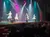 A group of performers is dancing on a stage with colorful spotlights, watched by an audience.
