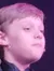 A young boy is performing on stage in a black suit with sparkly details and a flower accessory, under colorful stage lights with a girl in the background.
