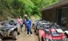 A group of people wearing helmets stand near a row of parked ATVs on a dirt path surrounded by lush green trees.