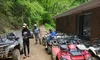 Several people are preparing for an off-road adventure with ATVs parked beside a brick building in a lush, forested area.