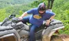 A person wearing a helmet and blue shirt is getting onto a muddy ATV in a scenic, wooded area.