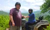 Two people wearing helmets are posing with an ATV on a dirt trail with a scenic, mountainous background.