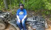 A person wearing a helmet and a blue T-shirt is sitting on an ATV in a wooded area.