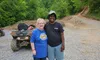 Two people are smiling and posing together on a gravel path with a forested background and an ATV nearby.