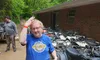 A person wearing a blue shirt and holding a helmet stands smiling next to several muddy ATVs in a wooded area.